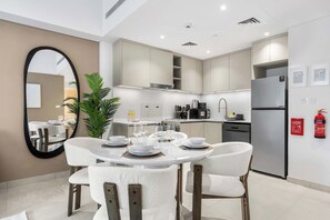 A modern kitchen with white cabinets, a stainless steel refrigerator, and a dining area with white chairs and a round table set for four. - StayShort - Modern 1BR Retreat in MJL Sleeps 4 (Madinat Jumeirah Living)