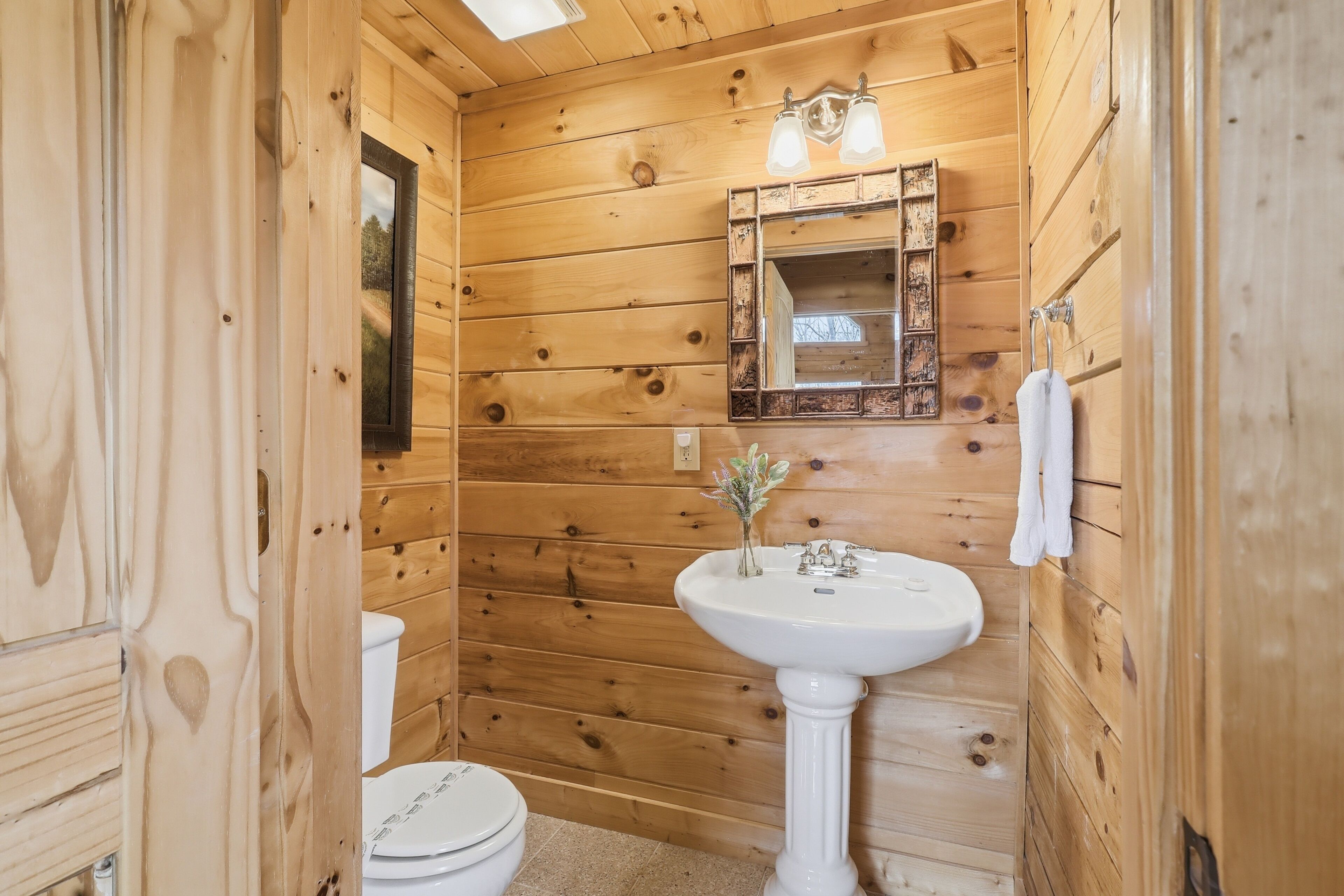 Un baño con paneles de madera, un lavamanos pedestal blanco, un espejo y un inodoro.