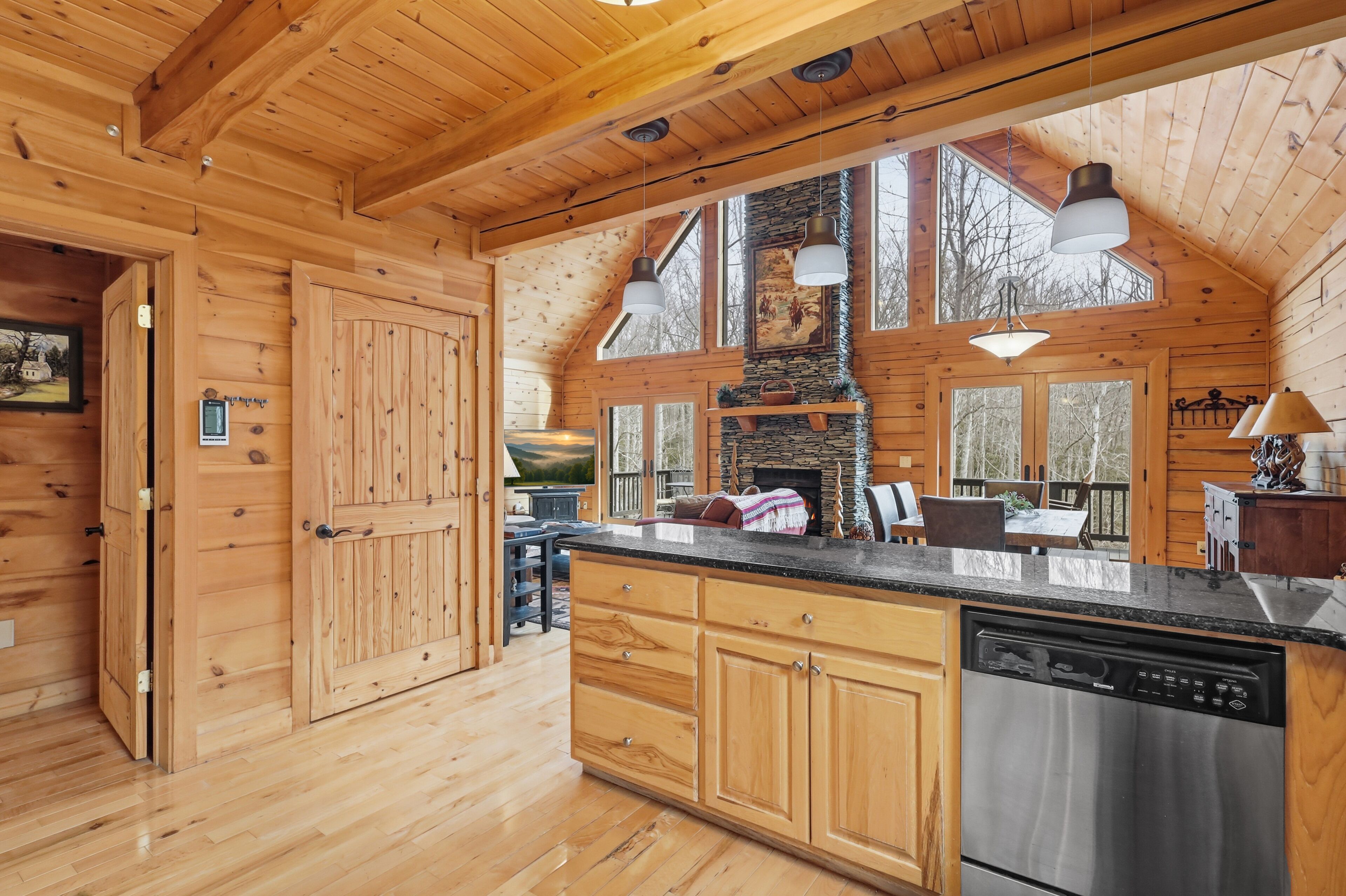 A wooden cabin interior with a kitchen, dining area, and a large window overlooking a deck.