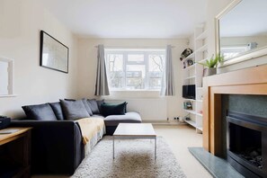 A living room with a sofa, a coffee table, a fireplace, a window with curtains, and a bookshelf. - Peaceful 2BDR Flat in Serene Clapham Cul de Sac (London)