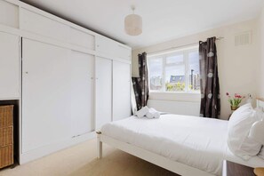 A bedroom with a bed, white wardrobe, window with curtains, and a view of buildings outside. - Peaceful 2BDR Flat in Serene Clapham Cul de Sac (London)