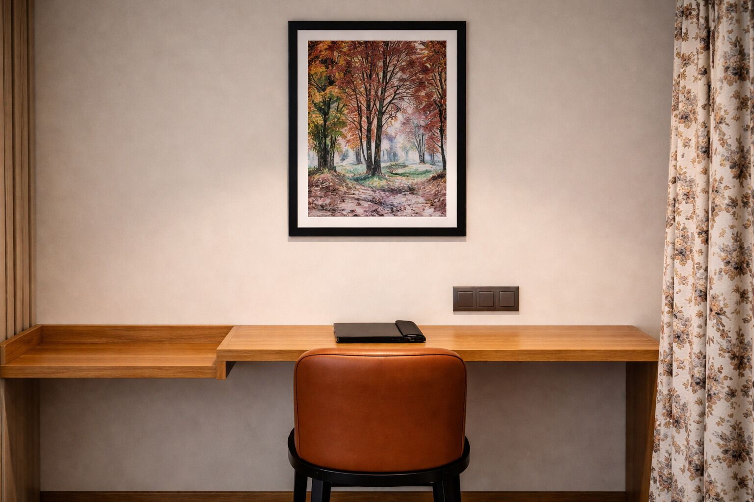 Un escritorio con una laptop, un cuadro enmarco de un bosque y una silla.