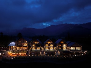 A row of wooden cabins with large windows, a sign for a restaurant, and a view of mountains in the background. - Mountnoor Sarovar Premiere Pahalgam (Anantnag)