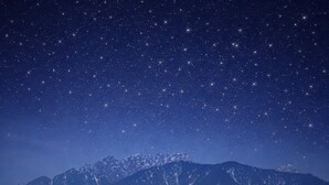 Un área de estar al aire libre con una mesa y sillas, una valla de madera y vistas a montañas nevadas bajo un cielo estrellado.