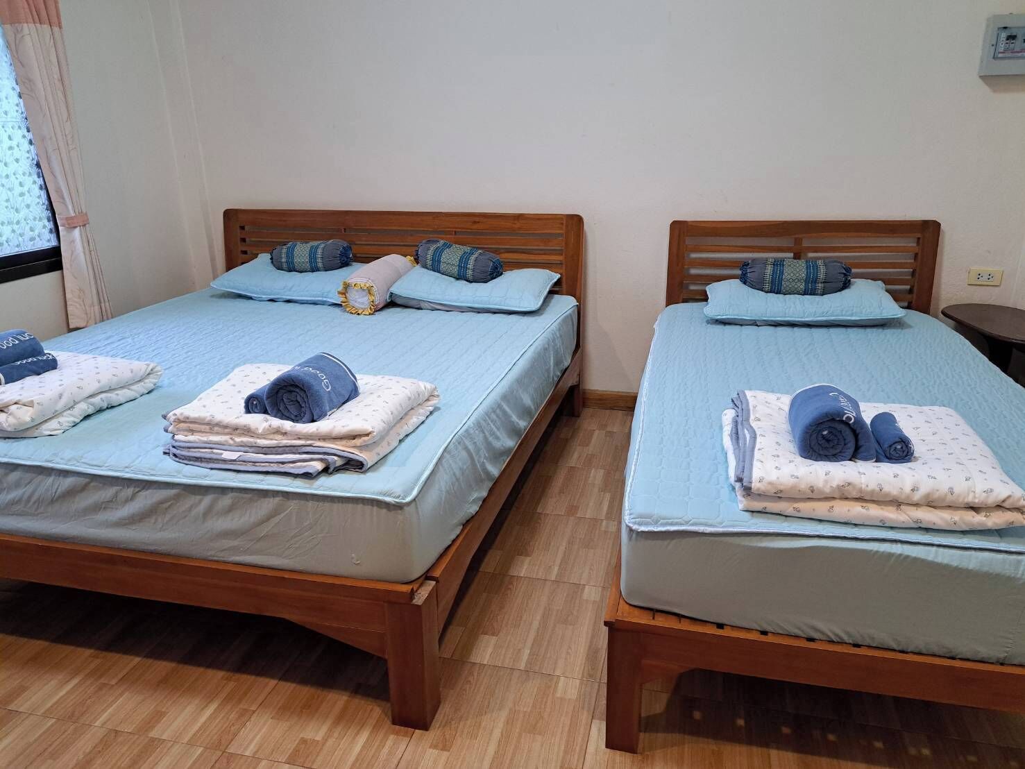 Duas camas de madeira com roupa de cama e almofadas azuis, um chão de madeira e uma janela com cortinas.