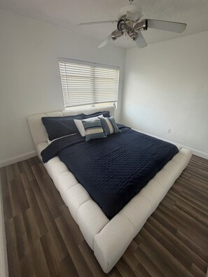Un dormitorio con cama, ventilador de techo, ventana con persianas y suelo de madera.