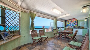 Ein überdachter Sitzbereich im Freien mit Holzstühlen und einem Tisch, Blick auf das Meer und einem farbenfrohen Wandbild.
