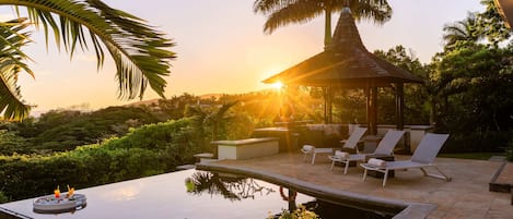A pool with lounge chairs and a gazebo, surrounded by greenery and palm trees.