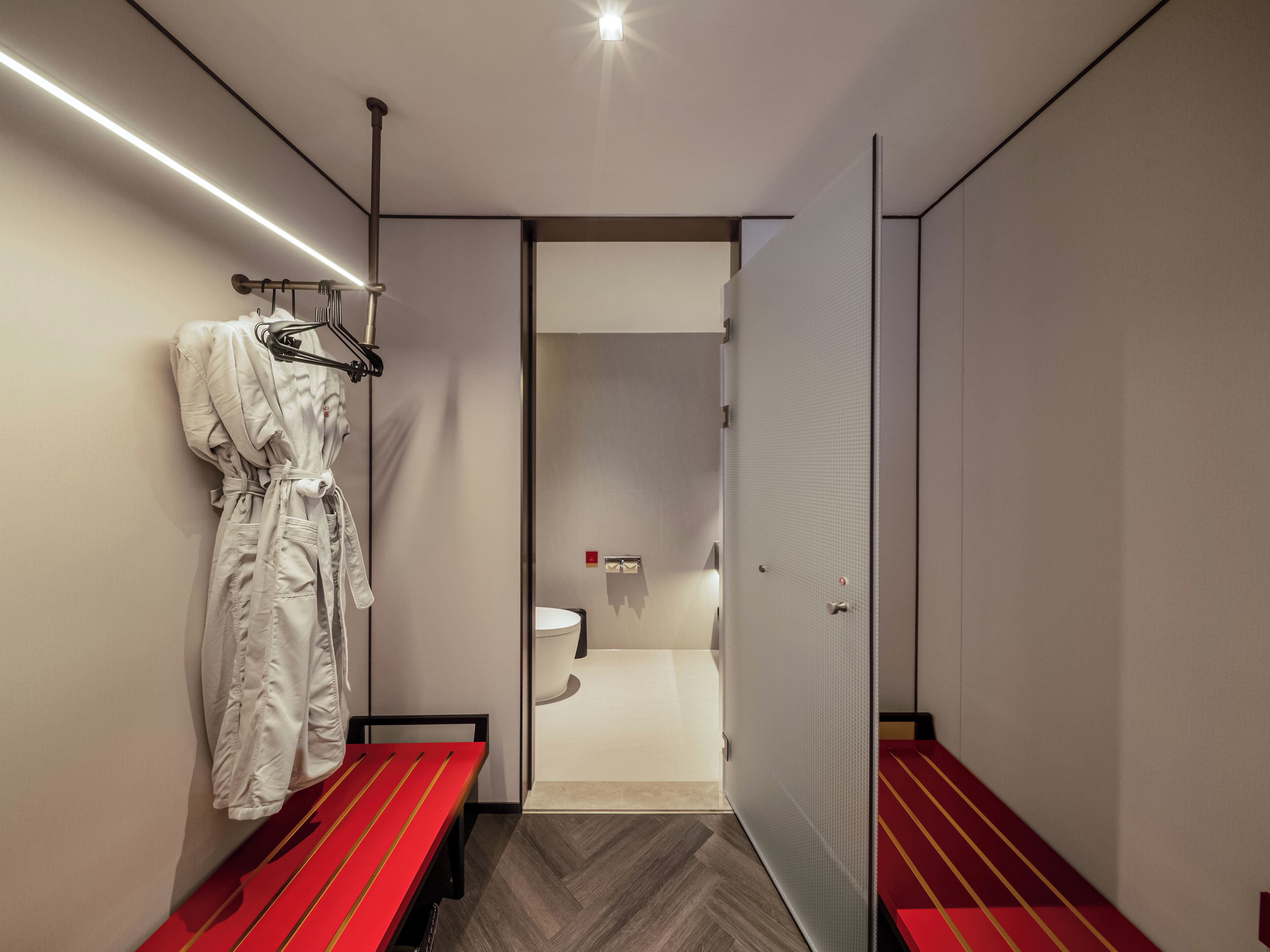 A hotel room with a bed, a bench, a robe hanging, and a toilet visible through an open door.