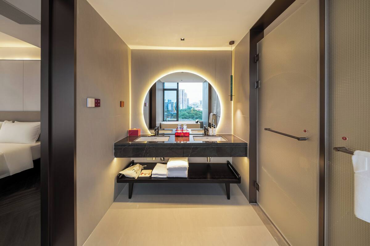 A modern hotel bathroom with a large mirror, dual sinks, a vanity, and a view of the city.