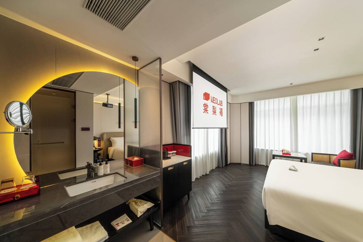 A hotel room with a large bed, a bathroom with a mirror and sink, a TV screen, and a large window with curtains.
