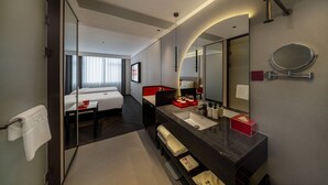 Habitació d'hotel moderna amb un llit gran, zona de seients separada, bany amb un mirall gran i un taulell amb productes de tocador.