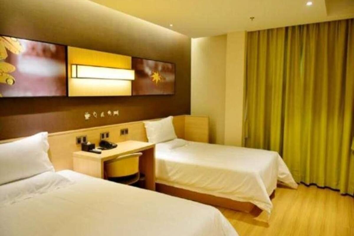 A hotel room with two beds, a desk, a nightstand, and a wall-mounted lamp.