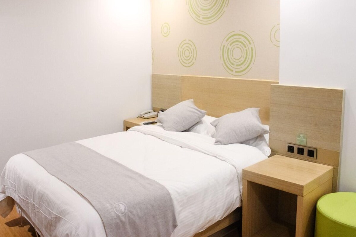 A neatly made bed with a wooden headboard, a bedside table with a lamp, and a green stool.