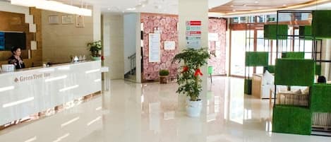 Zona de recepción de hotel con mostrador, una planta en maceta y un área de descanso con cojines verdes.