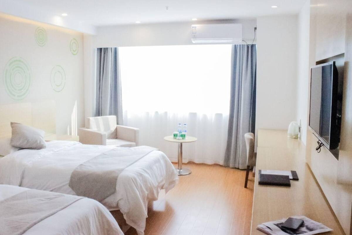 A hotel room with two beds, a TV, a small table with water bottles, and a window with curtains.