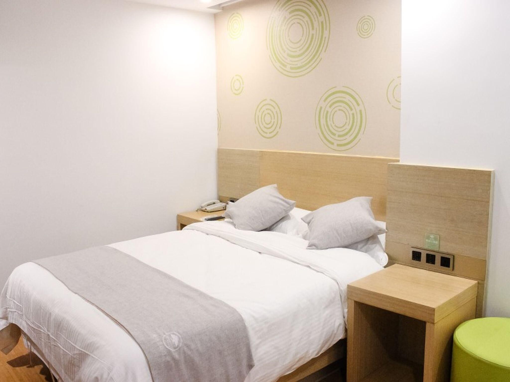 A neatly made bed with a wooden headboard, a bedside table with a lamp, and a green stool.