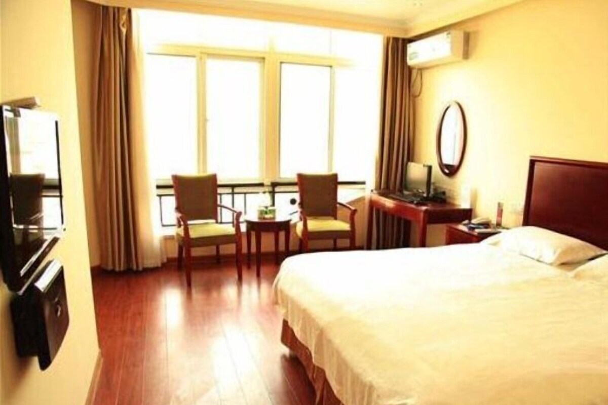 Quarto de hotel com cama, escrivaninha, duas cadeiras, televisão e janela com cortinas.