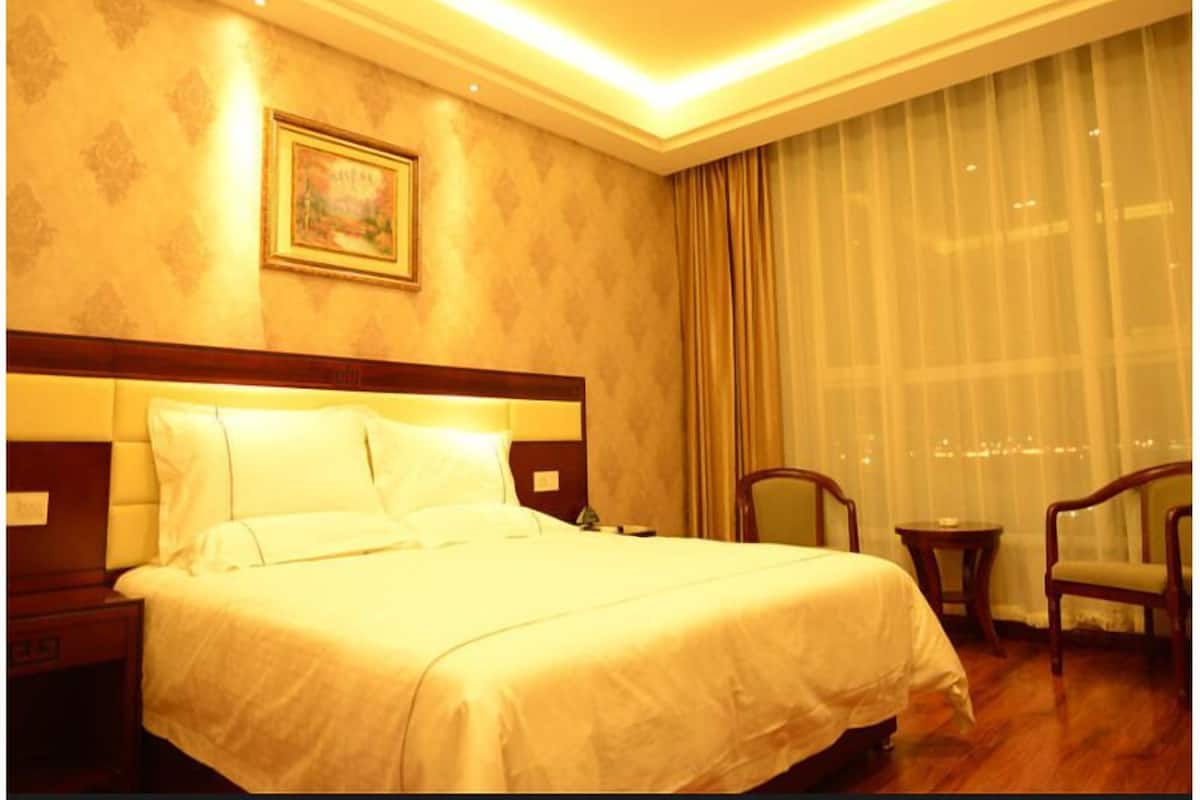 A hotel room with a large bed, a headboard, a nightstand, a chair, a small table, and a window with curtains.