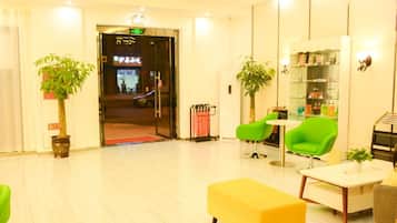 A hotel lobby with a reception desk, green chairs, a yellow sofa, and potted plants.
