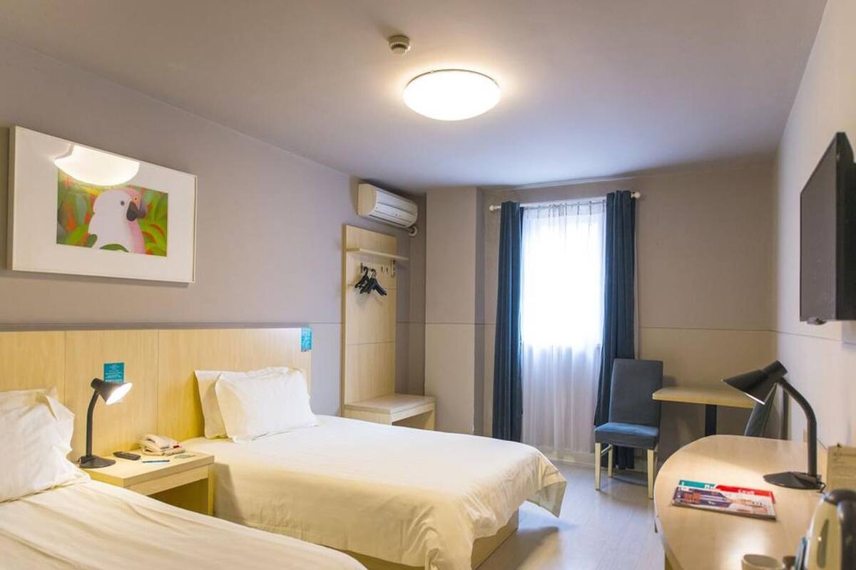 Hotellihuone, jossa on kaksi sänkyä, työpöytä, tuoli, ilmastointilaite ja ikkuna, jossa on verhot.