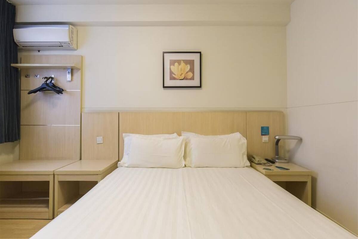 Hotellihuone, jossa on sänky, yöpöytä, seinälle ripustettava naulakko, seinään asennettu ilmastointilaite ja kehystetty taulu.