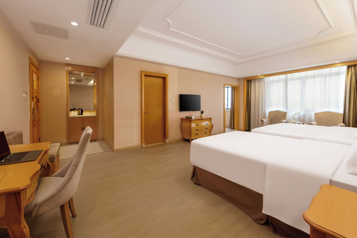 Quarto de hotel com cama, escrivaninha com laptop, televisão e área de estar com cadeiras.