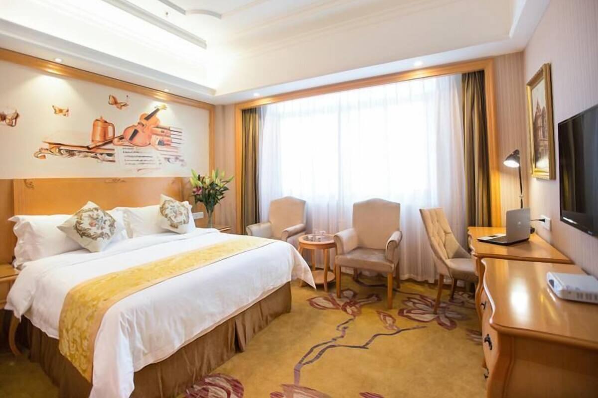 A hotel room with a large bed, two armchairs, a desk with a laptop, and a TV.