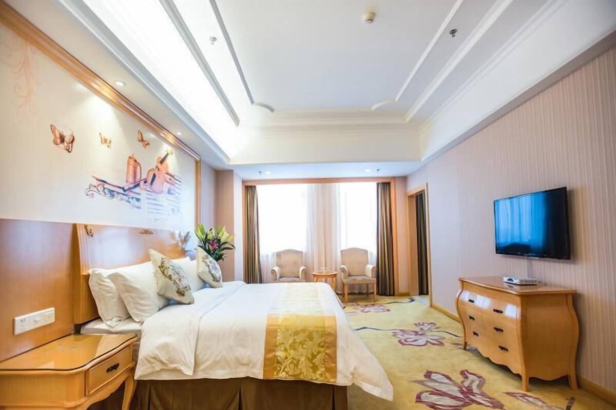 A hotel room with a bed, bedside tables, a TV, and a view of the outside.
