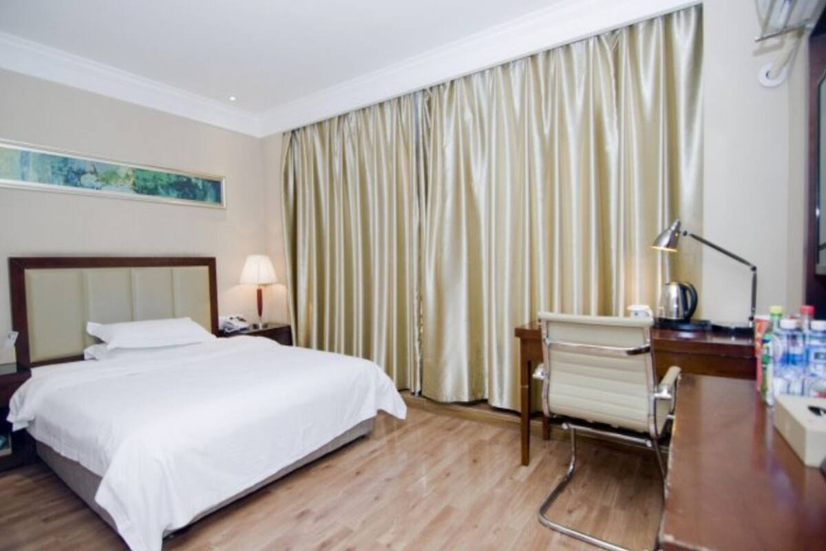 A hotel room with a bed, a desk, a chair, a lamp, and a curtain.