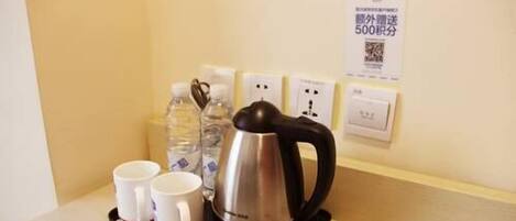 A kettle, two cups, and a water bottle on a table with a power outlet and a sign in the background.