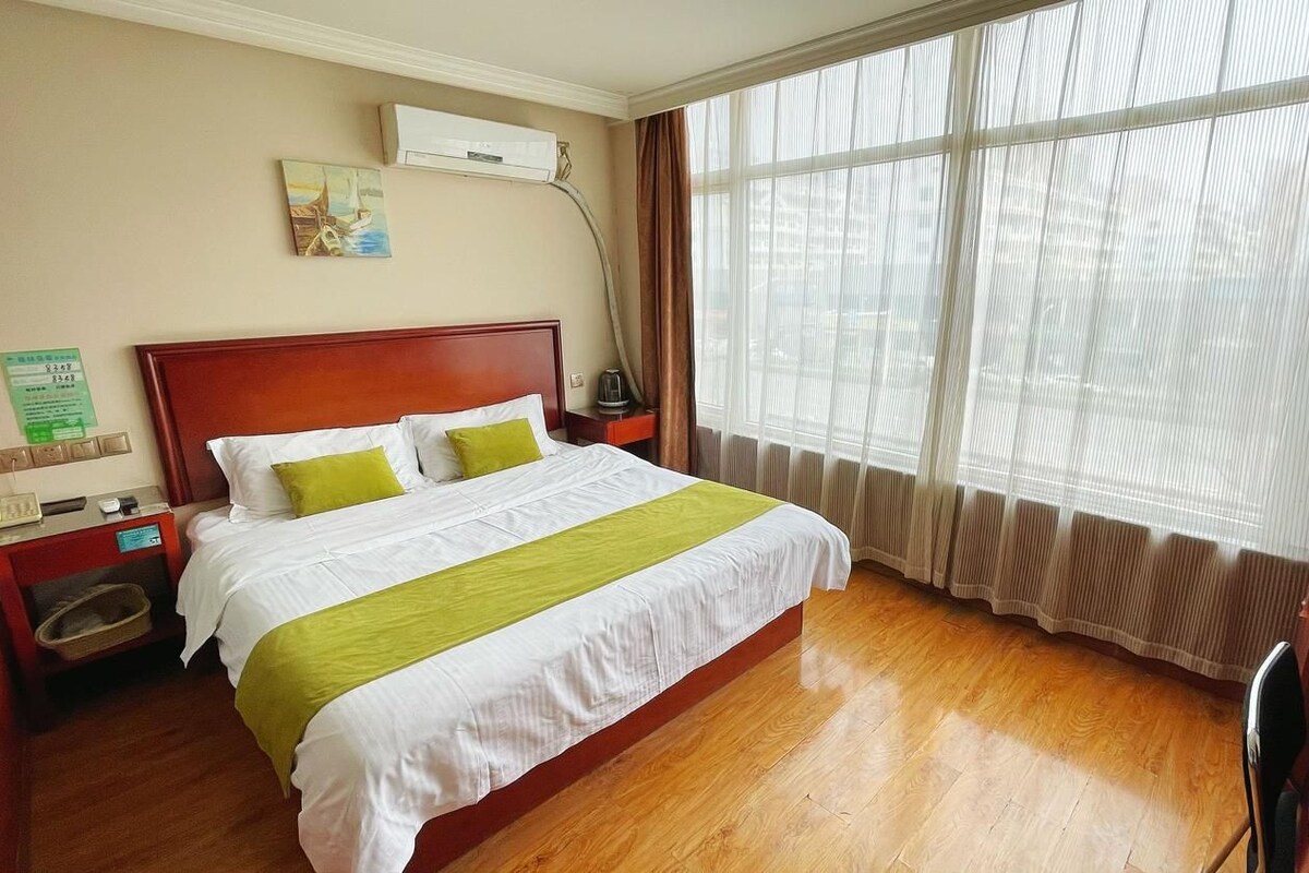 Una habitación de hotel con piso de madera, cama con ropa de cama blanca y verde, mesita de noche, lámpara y ventana con cortinas.