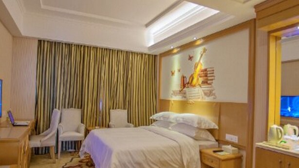 A hotel room with a bed, two chairs, a desk, a nightstand, and a wall with a guitar painting. - Vienna Classic Hotel Anhui Tongcheng (Anqing)