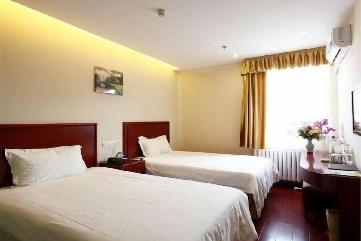 Habitació d'hotel amb dos llits individuals, una taula de nit amb una làmpada, una finestra amb cortines i un aire condicionat fixat a la paret.