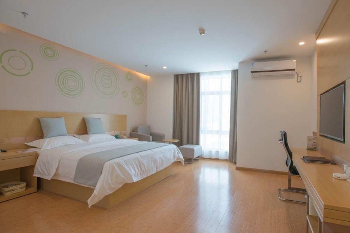A modern hotel room with a large bed, bedside tables, a desk with a chair, a TV, and a window with curtains.