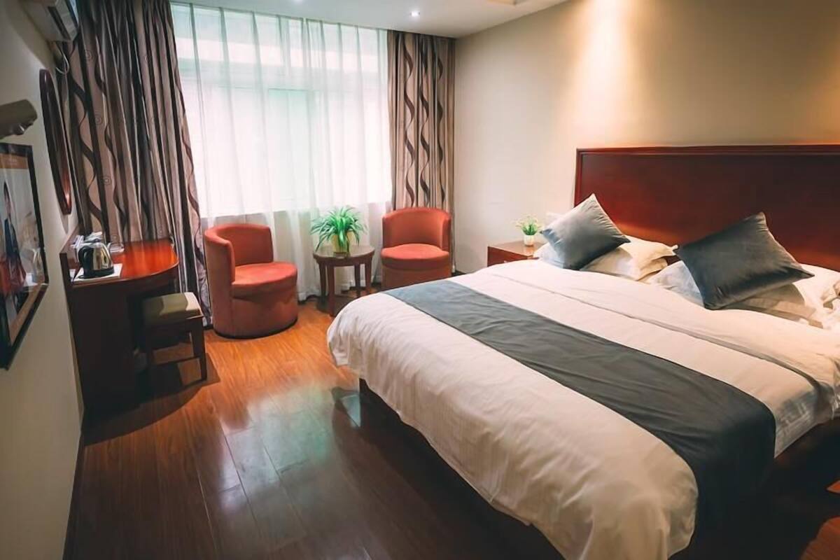 Une chambre d’hôtel avec un grand lit, deux fauteuils rouges, une petite table avec une plante et une vue sur l’extérieur à travers des rideaux légers.