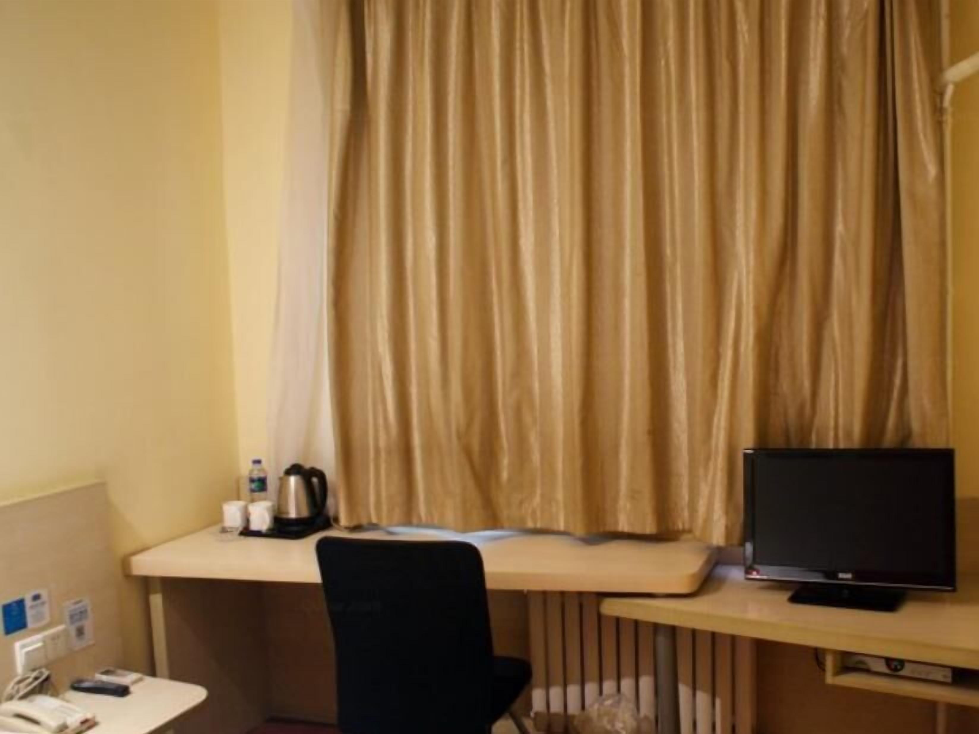 A hotel room with a desk, a chair, a television, a curtain, and a small table with a kettle and a cup.