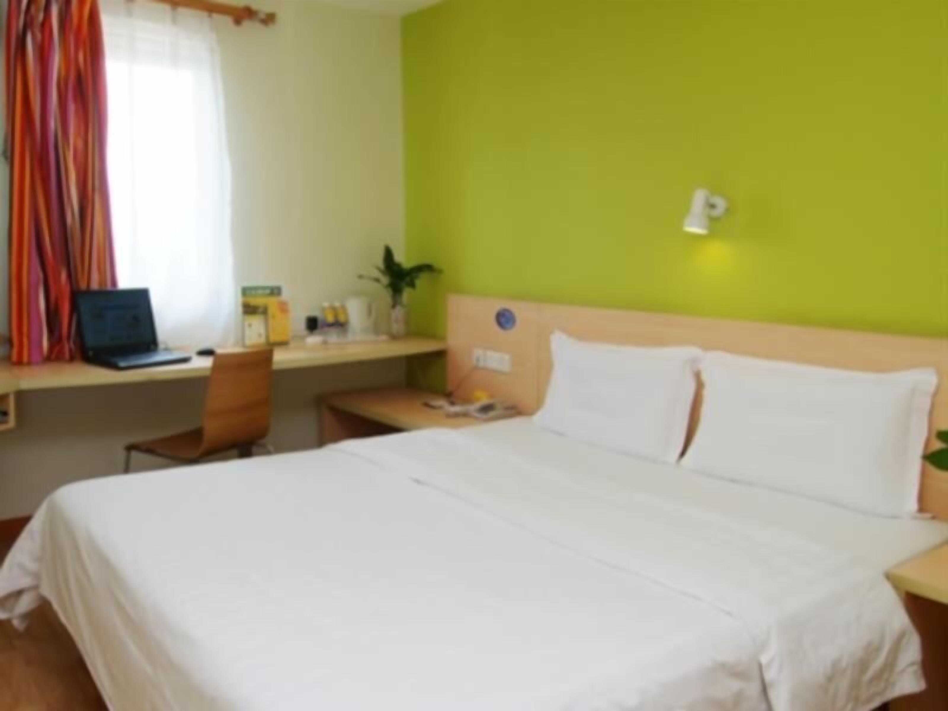 A hotel room with a bed, a desk with a laptop, a chair, and a small table with a plant.