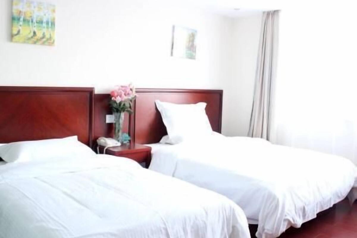 A hotel room with two single beds, a wooden headboard, a bedside table with a vase of flowers, and a painting on the wall.