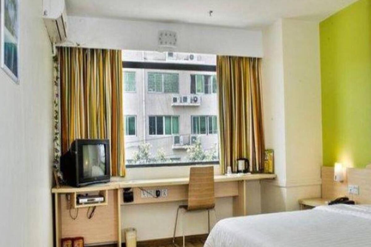 Habitación de hotel con cama, escritorio con televisor, una silla, ventana con cortinas y vista a edificios.
