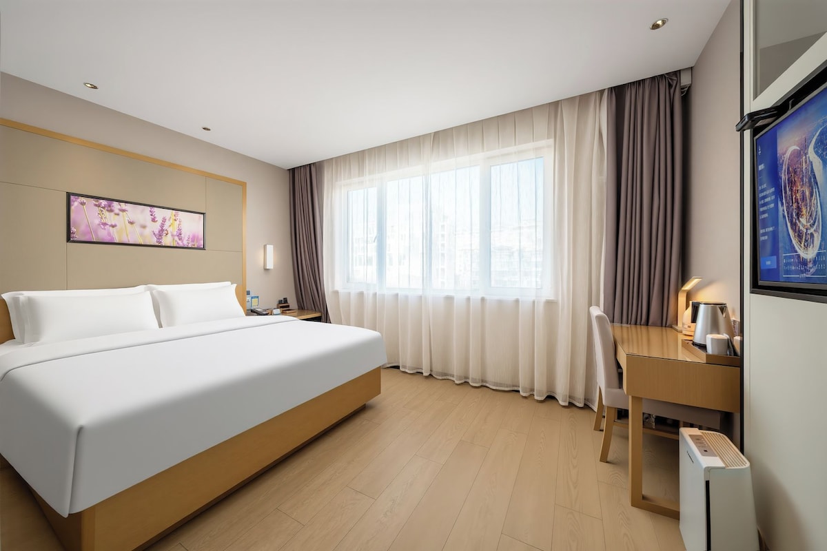 Ein Hotelzimmer mit einem großen Bett, einem Schreibtisch, einem Sessel, einem Flachbildfernseher und einem Fenster mit Vorhängen.