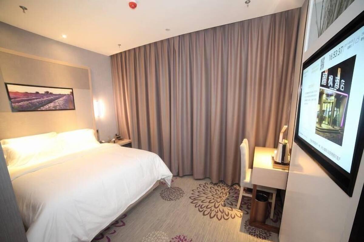 Ein Hotelzimmer mit einem Bett, einem Schreibtisch, einem Stuhl, einem großen Fenster mit Vorhängen, einem an der Wand befestigten Fernseher und einem Teppich mit Blumenmuster.