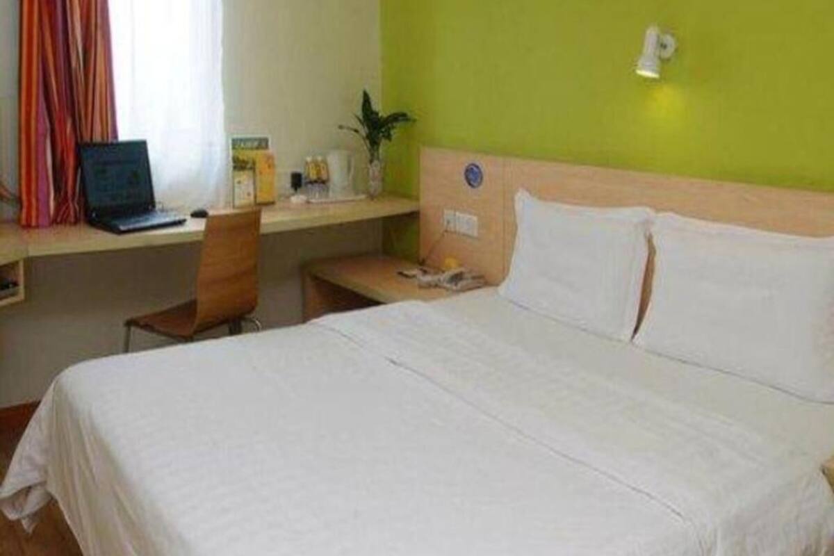 Una habitación de hotel con cama, escritorio con una computadora portátil, silla, mesita de noche y lámpara de pared.