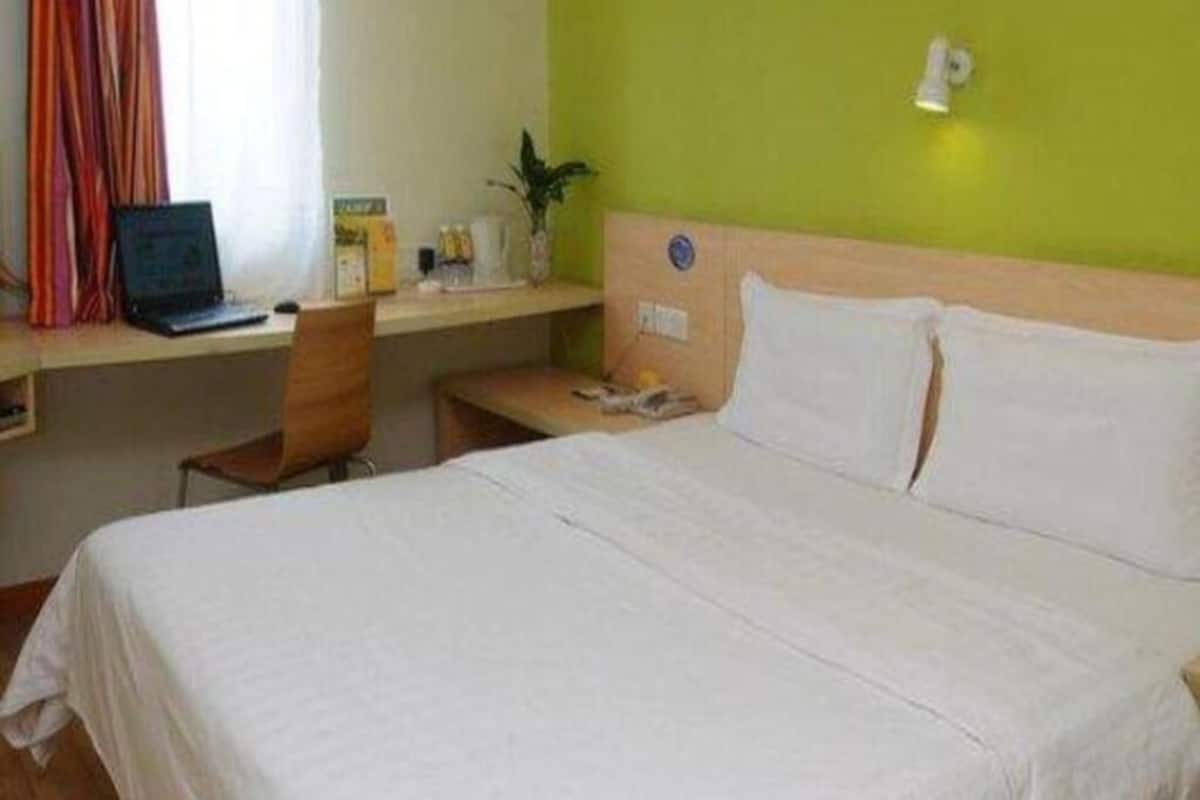 A hotel room with a bed, a desk with a laptop, a chair, a nightstand, and a wall-mounted lamp.
