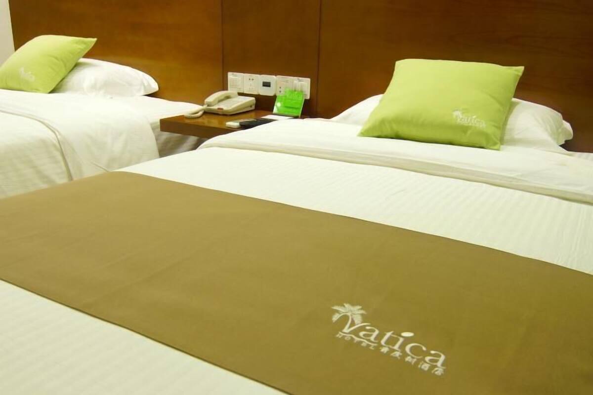 Ein Hotelzimmer mit zwei Betten, jeweils mit weißer Bettwäsche und grünen Kissen, einem Holz-Kopfteil, einem Nachttisch mit Telefon und einem grünen Gegenstand sowie einer braunen Matte mit dem Schriftzug 'Vatica'.