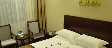A hotel room with a bed, two chairs, a nightstand, a painting, and a window with curtains.