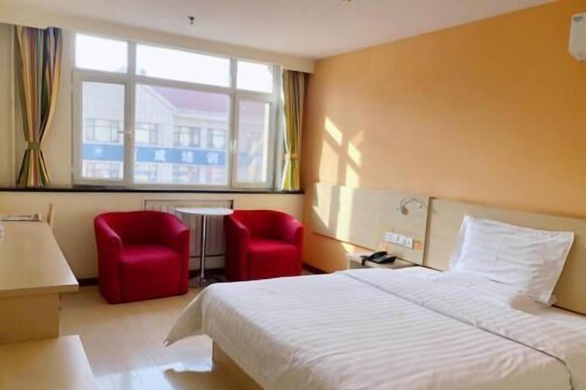 A hotel room with a bed, two red chairs, a small table, and a window with curtains.