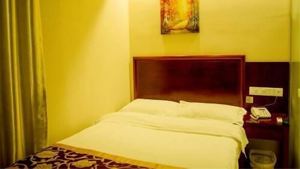 A hotel room with a bed, a painting on the wall, and a nightstand with a phone. - Greentree (Fuyang)