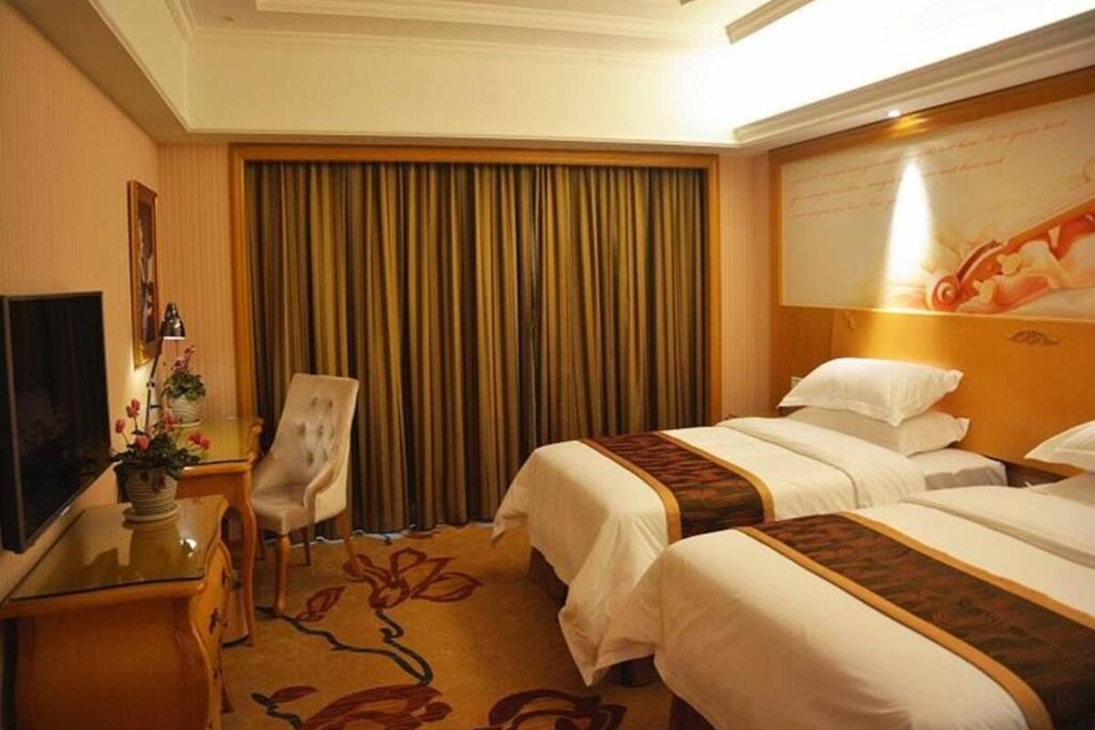 A hotel room with two beds, a desk, a chair, a television, and a large window with curtains.