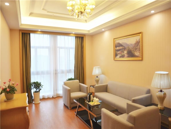 Ein Hotelzimmer mit einer Couch, Sesseln, einem Couchtisch, einer Lampe, einem Gemälde und einem Fenster mit Vorhängen.